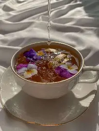 Une tasse de thé blanche avec des fleurs de violettes et blanches et des tranches de fruits, avec de l'eau versé délicatement à l'intérieur, posée sur un drap blanc plissé.